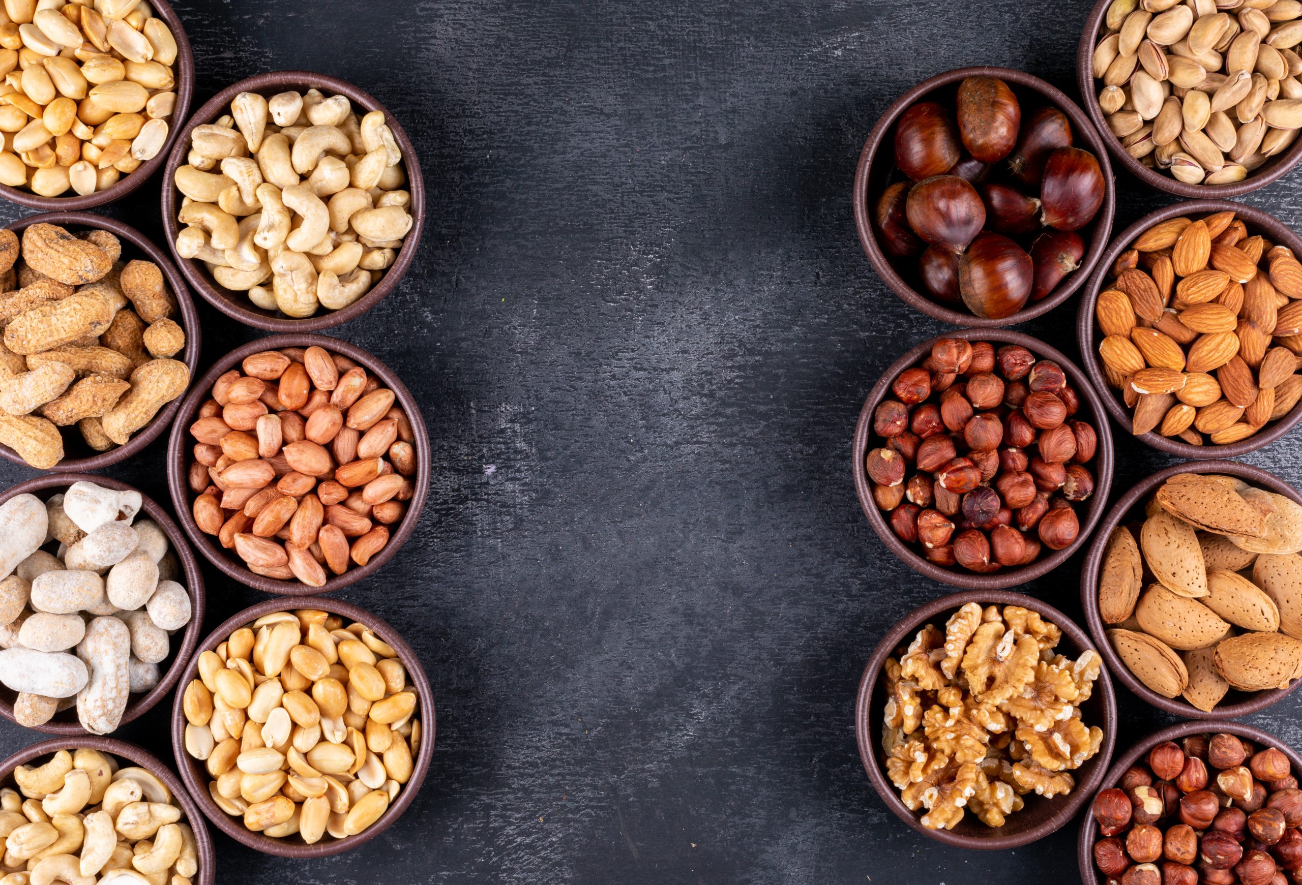 Flat lay assorted nuts and dried fruits in mini different bowls with pecan, pistachios, almond, peanut, on black stone background. horizontal free space for your text